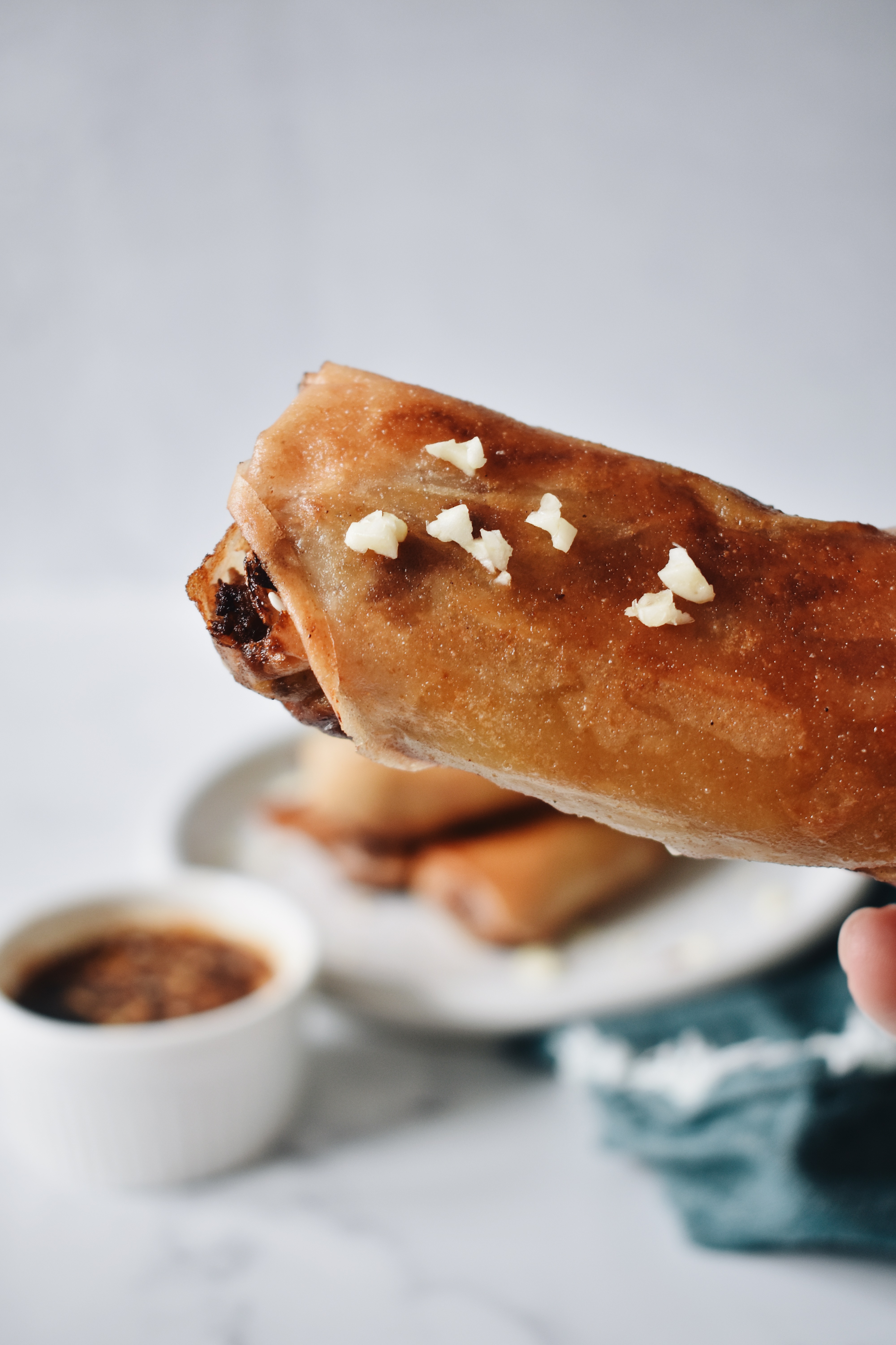 filipino spring roll with some garlic pieces on it help up to them camera with sauce, plate of lumpia, and napkin in the background.