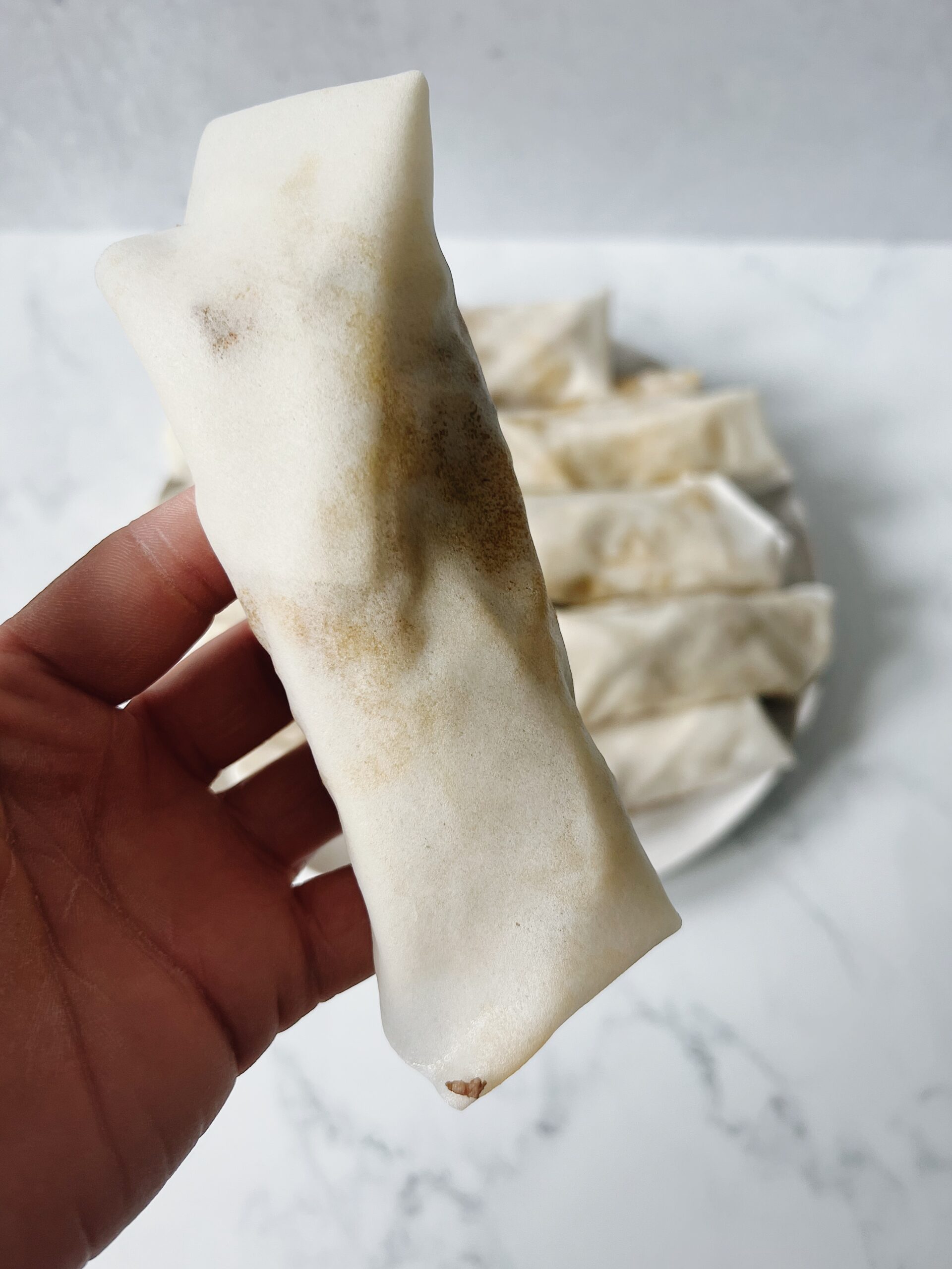 mean and filling wrapped in spring roll wrapper and held up to the camera with other rolled lumpia on a plate in the background.