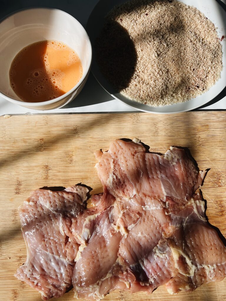 raw chicken sitting on the cutting board pounded with panko and eggwash on the side.