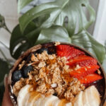 Acai bowl with bananas, strawberries, blueberries, granola, and honey in Coconut bowl with monstera leaves in the background