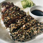 Furikake ahi cooked and sitting on plate with rice, avocado, and shoyu vinegar sauce.